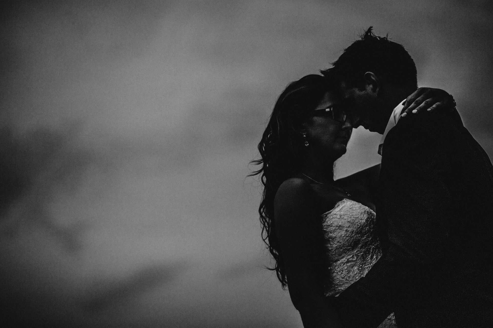 Czarno-białe zdjęcie pary obejmującej się na tle pochmurnego nieba. Panna młoda w koronkowej sukni i okularach przeciwsłonecznych, pan młody w ciemnym garniturze.