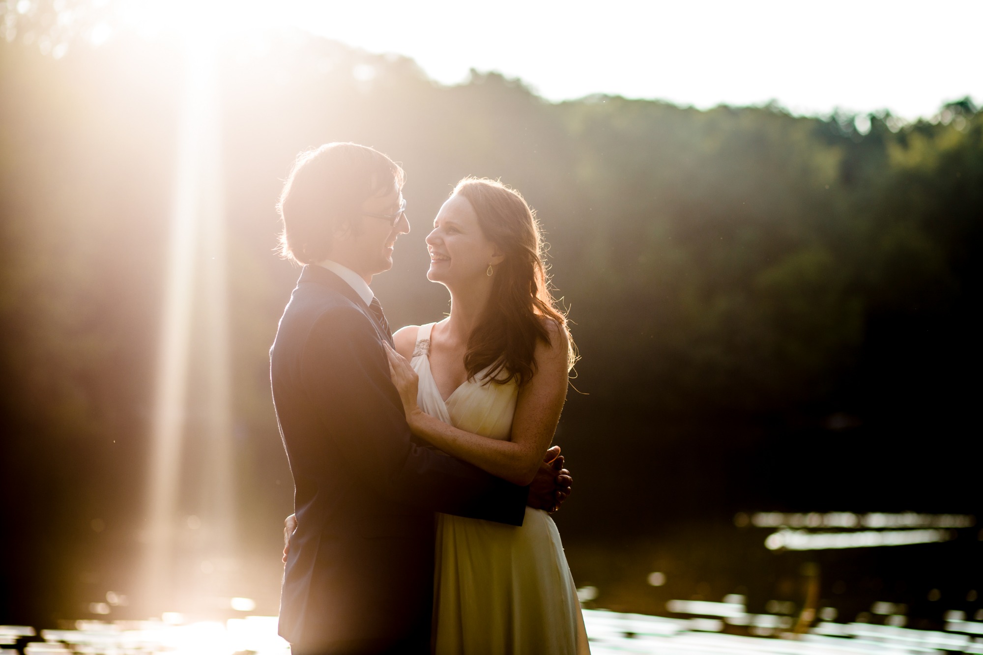 Para młoda w objęciach na tle jeziora w promieniach zachodzącego słońca, kobieta w zwiewnej sukni, mężczyzna w garniturze, oboje uśmiechnięci.