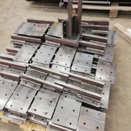 Stos metalowych płyt z otworami i przyspawanymi profilami, ułożonych na palecie, widoczne ślady po spawaniu.