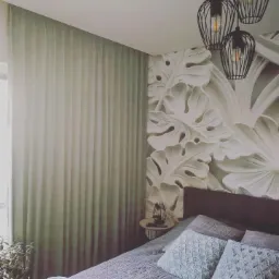 Jasne, gładkie zasłony w sypialni z łóżkiem w odcieniach szarości i błękitu, dekoracyjna ściana z motywem liści monstery i wiszące lampy w stylu industrialnym.