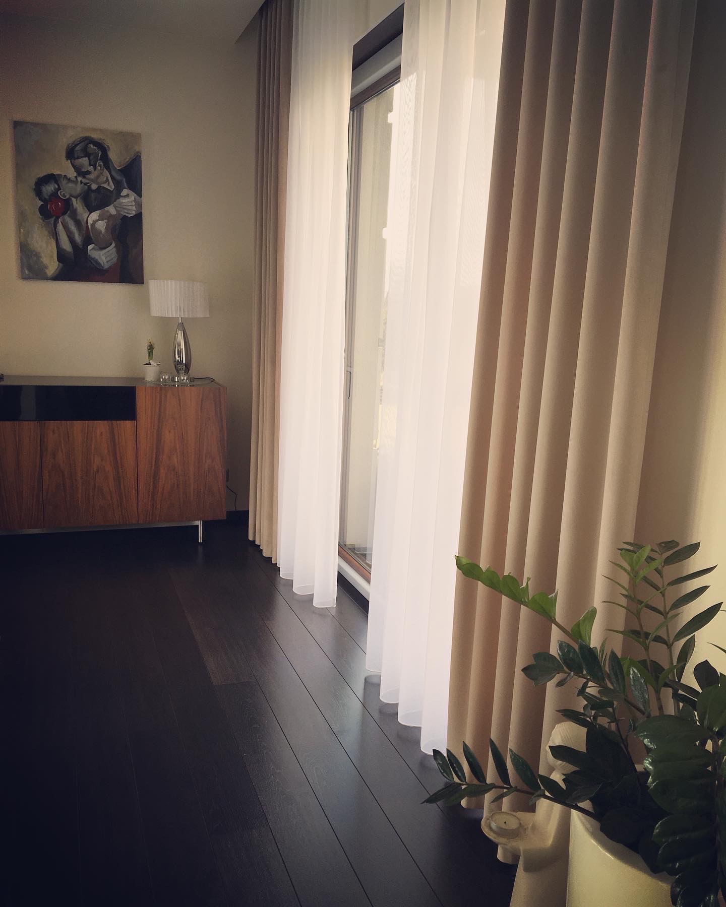 Beżowe zasłony zaciemniające i białe firany przy oknie balkonowym, uzupełnione obrazem na ścianie, drewnianą komodą i doniczkową rośliną na pierwszym planie.