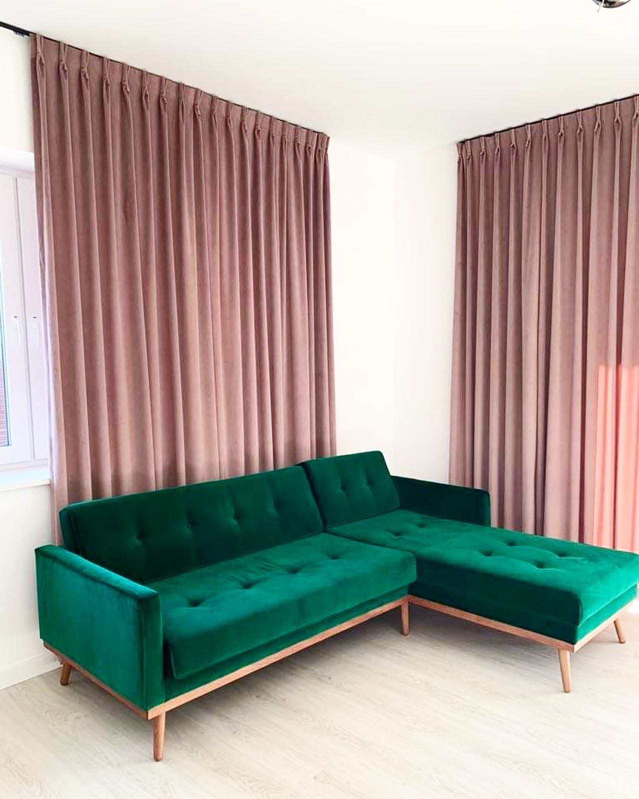 Jasnoróżowe, długie zasłony panelowe na szynach sufitowych, po bokach okna, w tle zielona, welurowa sofa narożna na drewnianych nóżkach.