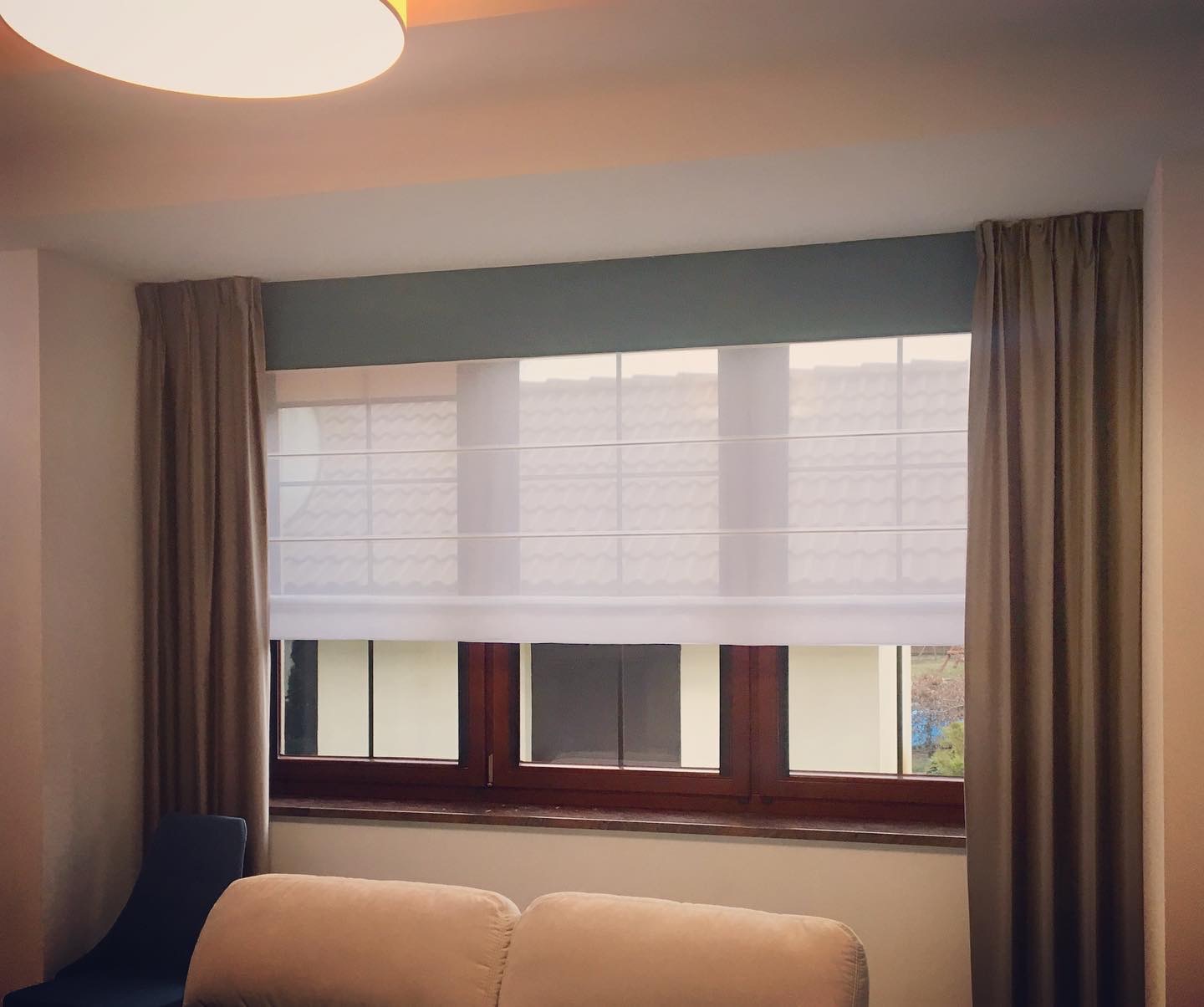 Okna z brązowymi ramami i białymi roletami rzymskimi oraz beżowymi zasłonami po bokach, widok z wnętrza pokoju, oświetlenie górne.