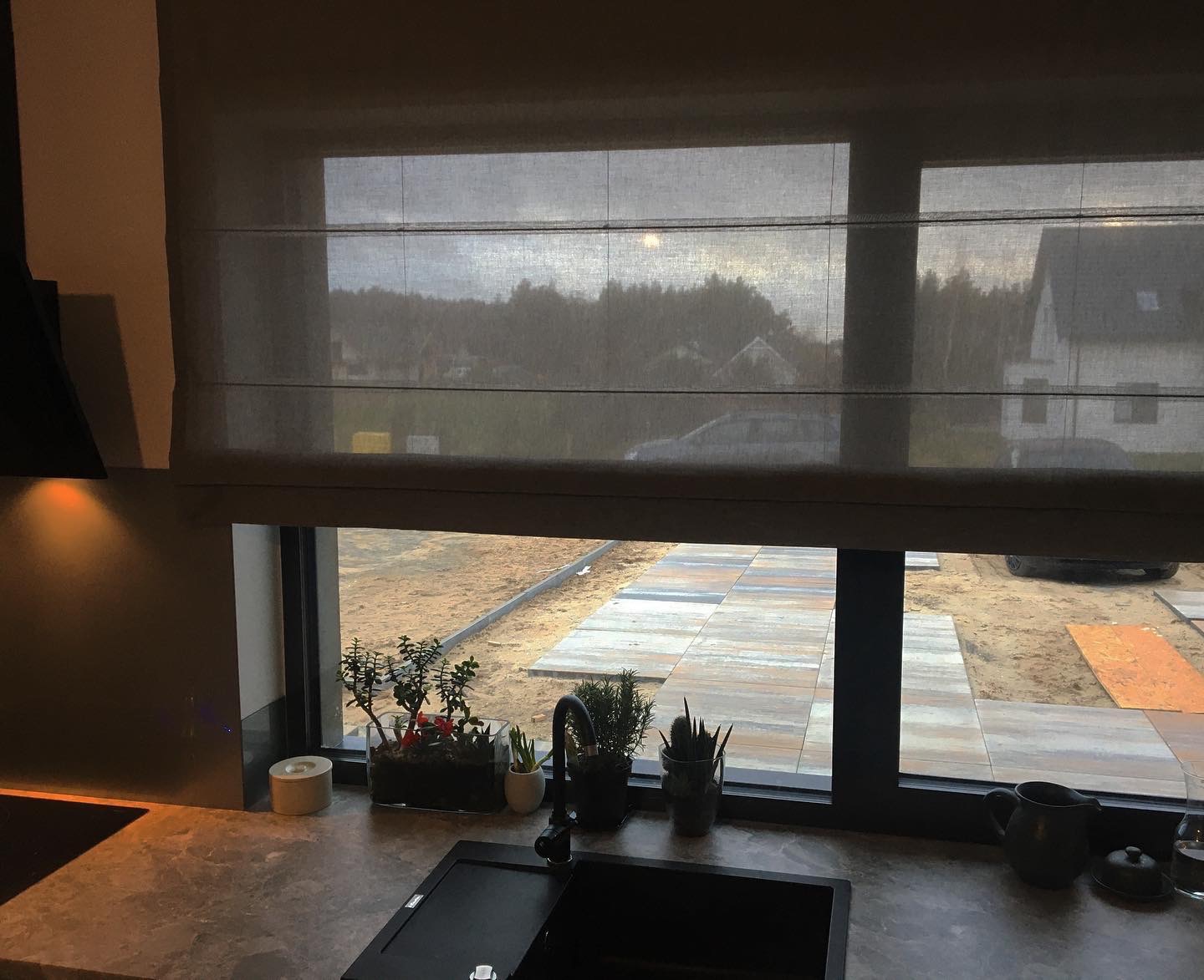 Szare zasłony rzymskie w oknie kuchennym z widokiem na budowę chodnika z kostki brukowej i dom w tle, rośliny doniczkowe na parapecie.