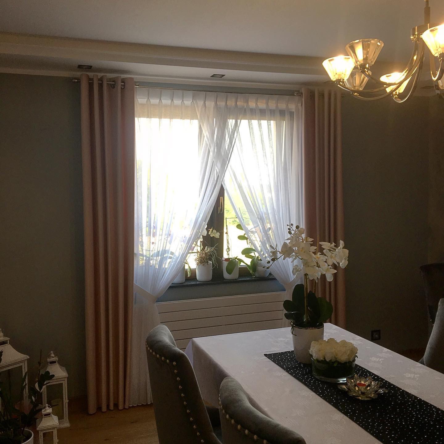 Eleganckie, pudrowo-różowe, zasłony po bokach okna z białymi, zwiewnymi firanami, doświetlonego dziennym światłem. Na parapecie doniczki z roślinami. Stół z białym obrusem i dekoracjami kwiatowymi...
