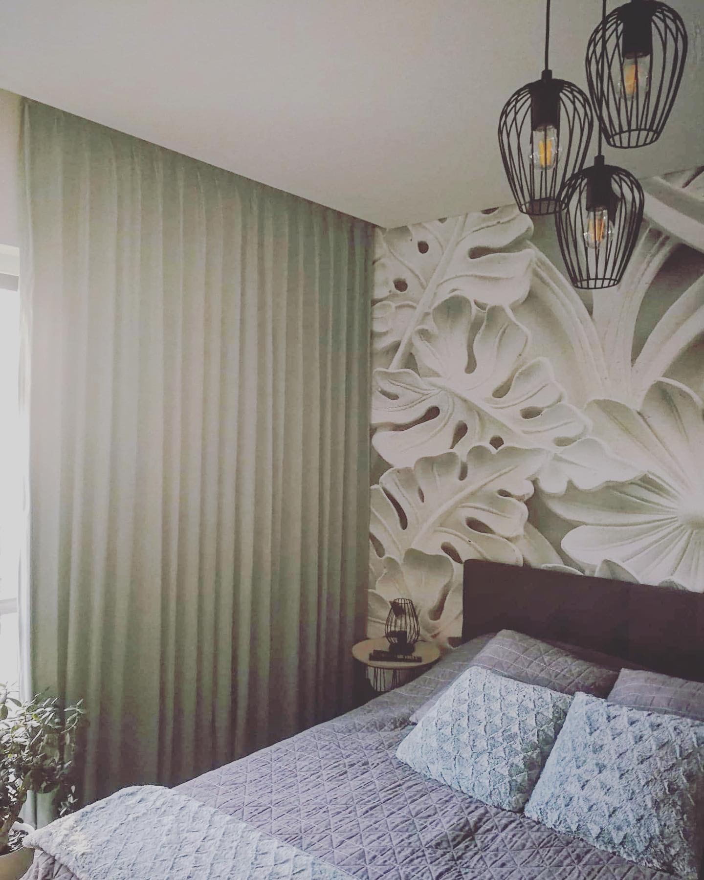 Jasne, gładkie zasłony w sypialni z łóżkiem w odcieniach szarości i błękitu, dekoracyjna ściana z motywem liści monstery i wiszące lampy w stylu industrialnym.