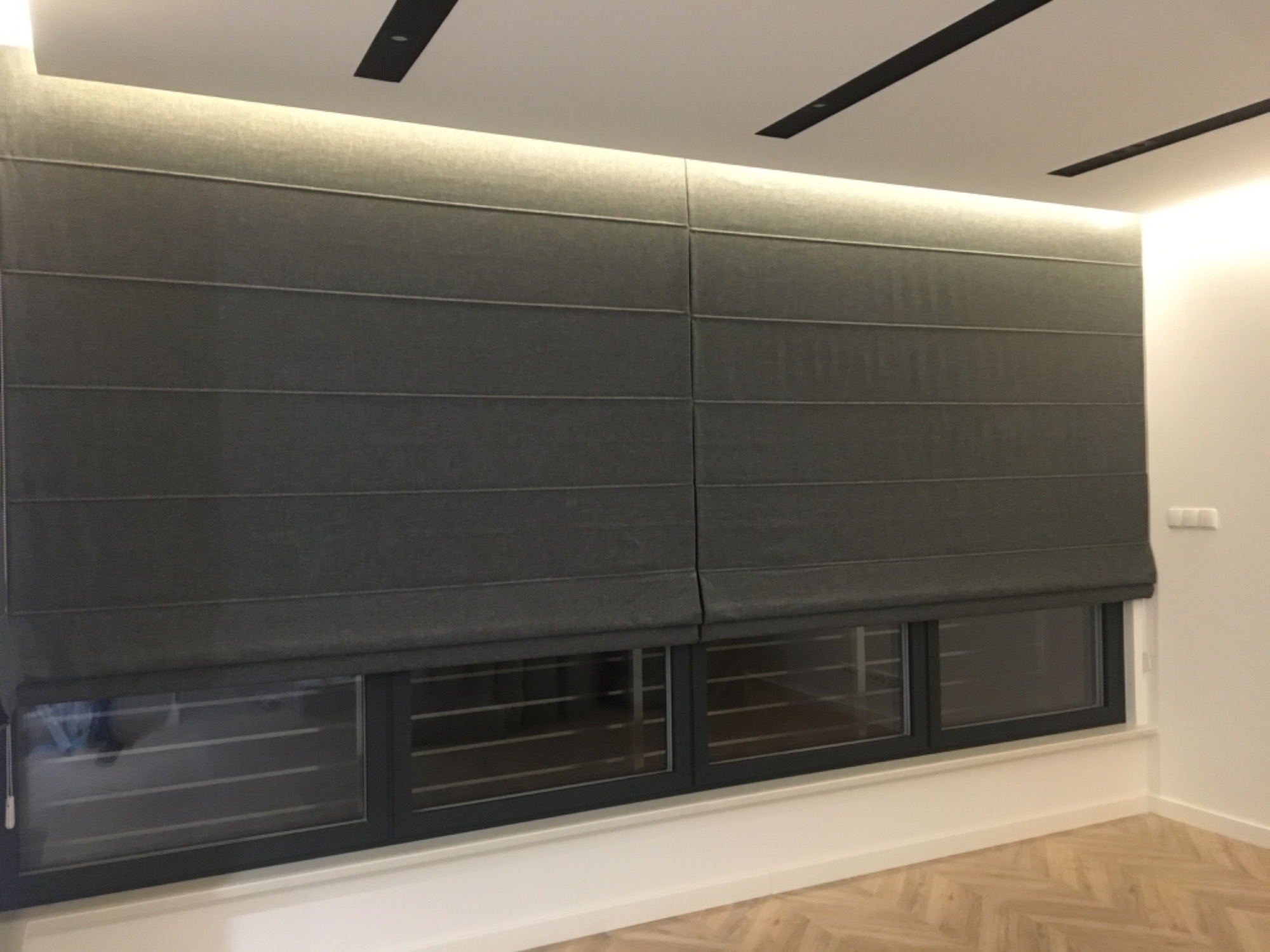 Szare, rzymskie rolety zaciemniające okna w nowoczesnym wnętrzu z drewnianą podłogą w jodełkę i oświetleniem LED w suficie.