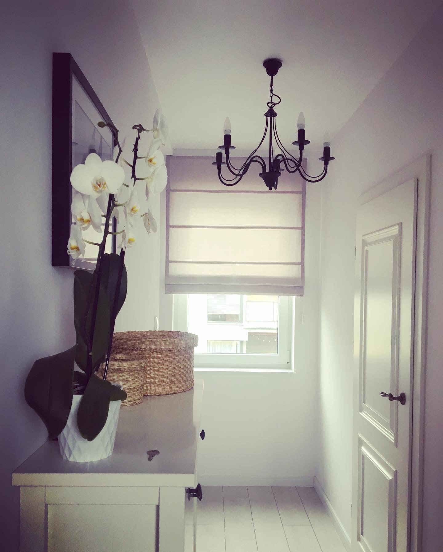 Jasne wnętrze: biała komoda z orchideą w doniczce, wiklinowe kosze, żaluzje rzymskie w oknie i czarny żyrandol.