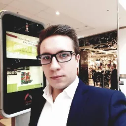 Mężczyzna w garniturze robi selfie na tle interaktywnego kiosku informacyjnego w galerii handlowej.