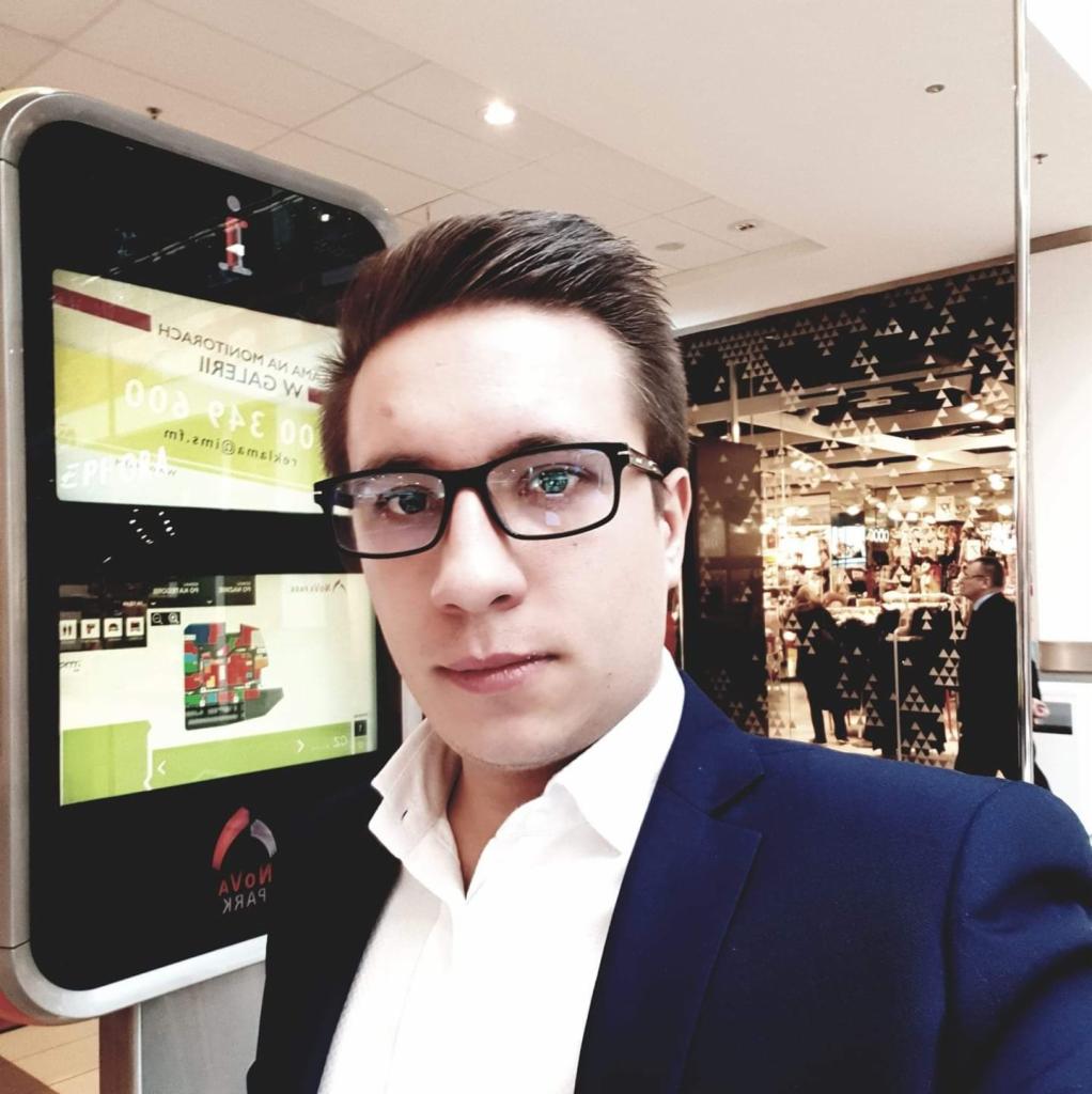 Mężczyzna w garniturze robi selfie na tle interaktywnego kiosku informacyjnego w galerii handlowej.