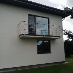 Balkon z prostą, metalową konstrukcją zabezpieczającą na tle białej ściany budynku i zielonego trawnika.