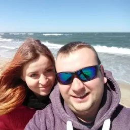 Selfie pary na plaży w słoneczny dzień; kobieta z rudymi włosami i mężczyzna w okularach przeciwsłonecznych, w tle wzburzone morze.