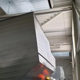 Montaż dużych, metalowych kanałów wentylacyjnych w hali przemysłowej; trzech pracowników w kamizelkach odblaskowych przytwierdza elementy konstrukcji do sufitu.