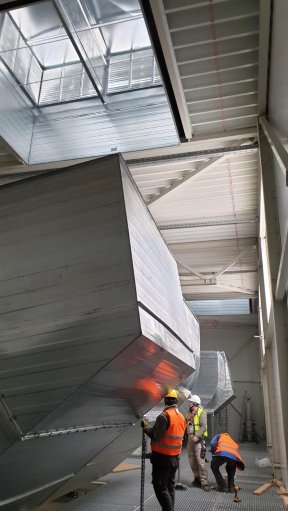 Montaż dużych, metalowych kanałów wentylacyjnych w hali przemysłowej; trzech pracowników w kamizelkach odblaskowych przytwierdza elementy konstrukcji do sufitu.
