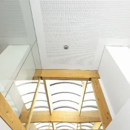 Sufit z perforowanej płyty, drewniana konstrukcja nośna z widocznymi śrubami, szklana balustrada i białe filary w nowoczesnym wnętrzu.