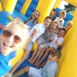 Radosna grupa dzieci w dmuchanym zamku zjeżdżalni, selfie z panią w okularach przeciwsłonecznych w Warce, jedna z dziewczynek ma pomalowaną twarz.