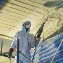 Pracownik w kombinezonie ochronnym natryskuje piankę PUR na sufit budynku, widoczne rusztowanie i plandeka ochronna.