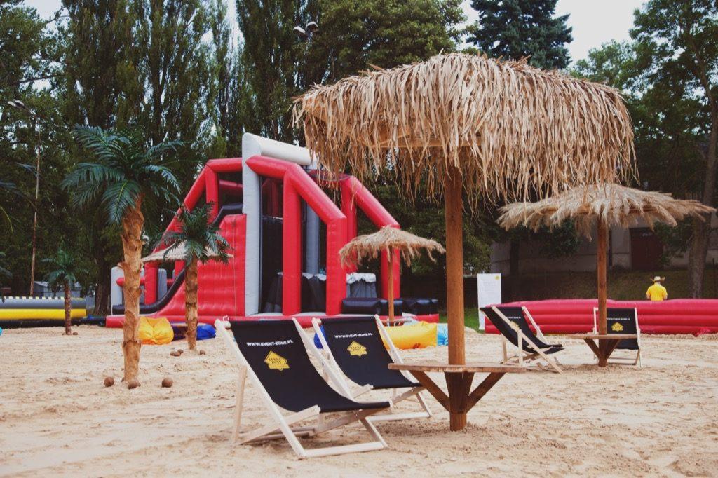 Plaża z dmuchanym zamkiem, palmami i leżakami z logo, tworząca strefę relaksu podczas eventu plenerowego.