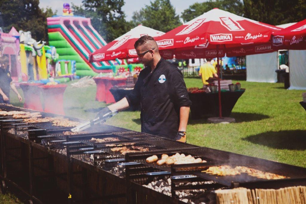 Kucharz w czarnym uniformie grilluje mięso i kiełbaski na długim grillu węglowym podczas imprezy plenerowej, w tle dmuchany zamek i parasole reklamowe.