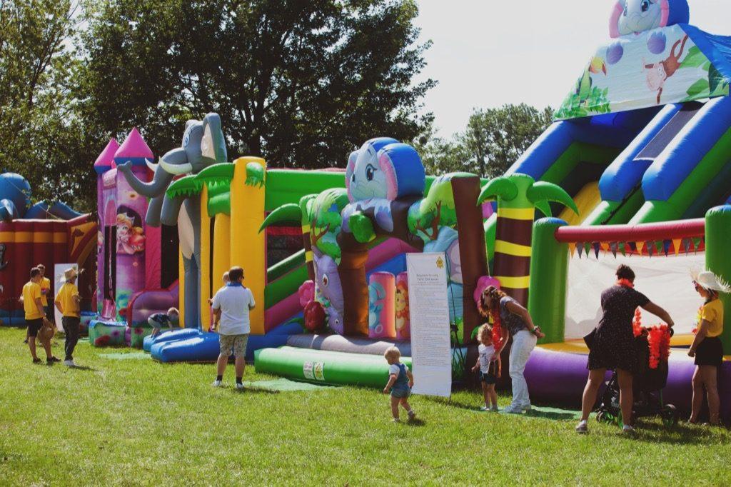 Rozległy park rozrywki z dmuchanymi zamkami i zjeżdżalniami o tematyce dżungli i zwierząt, z dziećmi bawiącymi się w pobliżu i dorosłymi nadzorującymi, na tle zielonej trawy i drzew.
