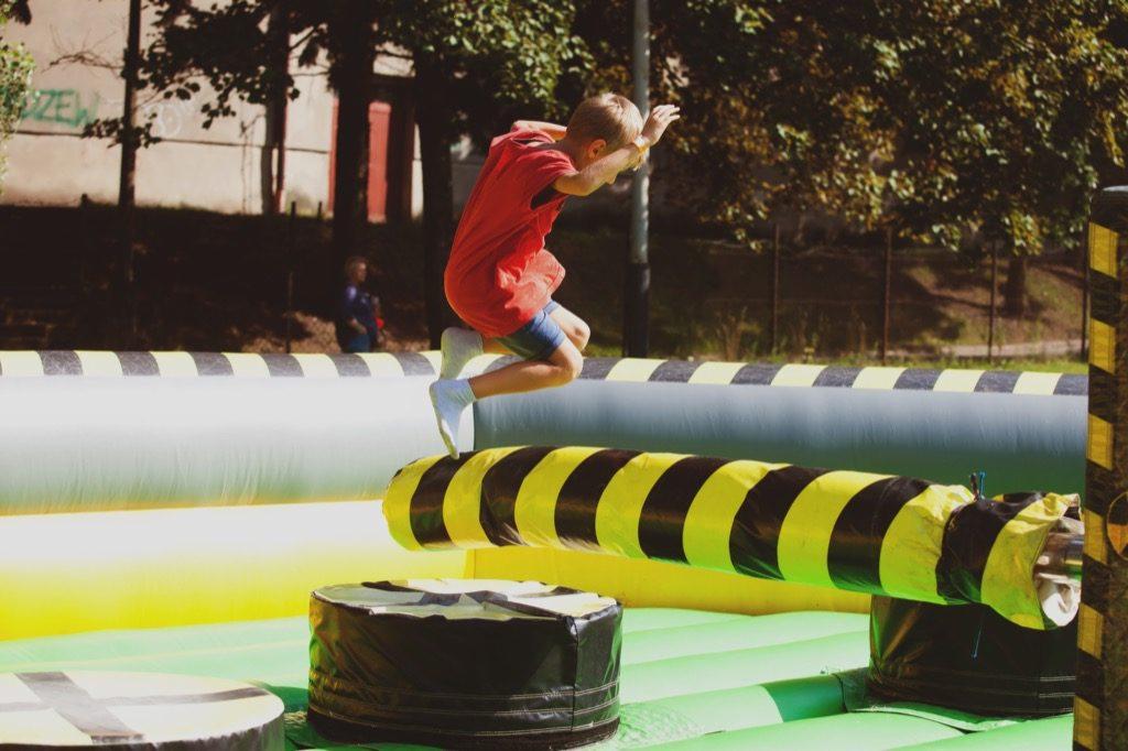 Chłopiec w czerwonej koszulce skacze na dmuchanym torze przeszkód z czarno-żółtymi pasami, w tle drzewa i ogrodzenie.