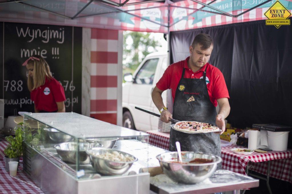 Mężczyzna w czerwonej koszulce i czarnym fartuchu z logo, trzyma pizzę na łopacie w stoisku z czerwono-białą kratą, obok widoczna kobieta i tablica z napisem 'Wynajmij mnie!!!', w tle biały samochód.
