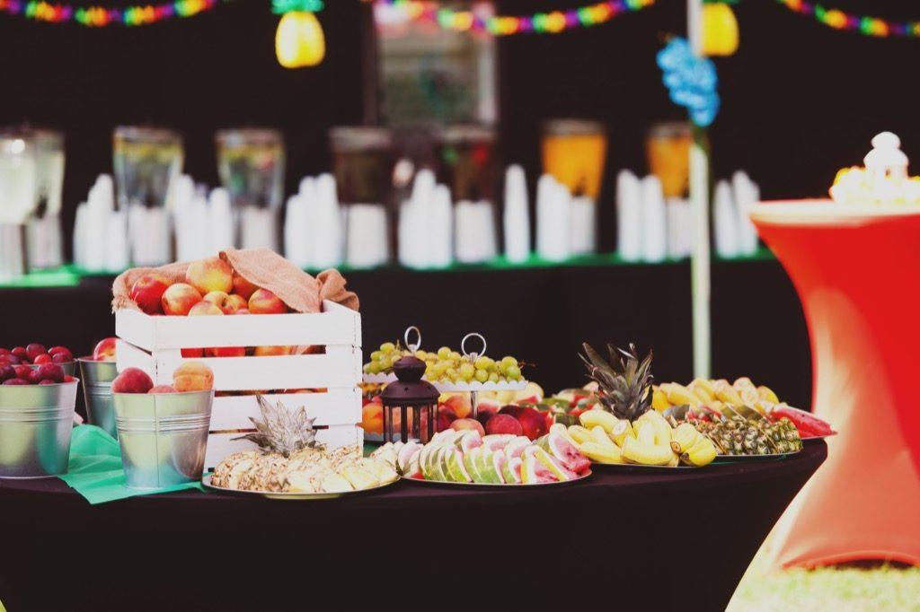 Stół z różnorodnymi owocami, arbuzem, ananasem, bananami, winogronami, brzoskwiniami i napojami w dyspenserach, udekorowany kolorowym łańcuchem świetlnym i lampionem, przygotowany na przyjęcie.