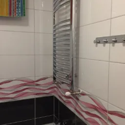 Chromowany grzejnik łazienkowy drabinkowy, zamontowany na ścianie z białych płytek z dekoracyjnym pasem bordowo-szarych fal, widoczna bateria wannowa i fragment wanny.