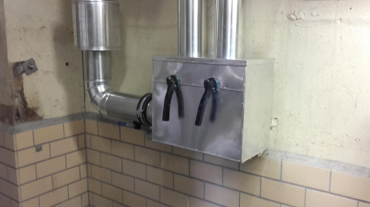 Fragment instalacji wentylacyjnej z metalowymi rurami i skrzynką, przymocowany do ściany wyłożonej częściowo cegłą, z widocznymi śladami wilgoci i niedoskonałościami tynku.