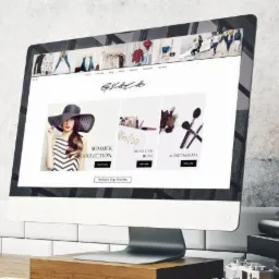 Ekran komputera iMac wyświetlający minimalistyczną stronę internetową z motywem modowym, ustawiony na drewnianym stoliku z książkami i staromodnym telefonem na tle ściany z efektem betonu.