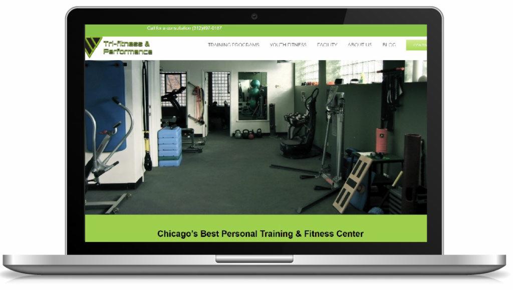 Laptop wyświetlający stronę internetową siłowni Tri-Fitness & Performance z widokiem na salę treningową z różnymi maszynami i sprzętem do ćwiczeń.