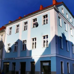 Odnowiona kamienica w Dzierżoniowie z błękitną elewacją, białymi pasami i oknami, widoczne detale architektoniczne, antena satelitarna na ścianie, sklep ogrodniczy w parterze.