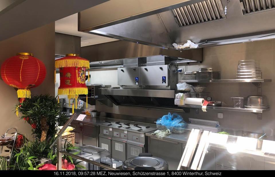 Profesjonalna kuchnia w azjatyckiej restauracji z systemem wentylacji nad frytkownicami, dekorowana czerwonymi lampionami i roślinami doniczkowymi.