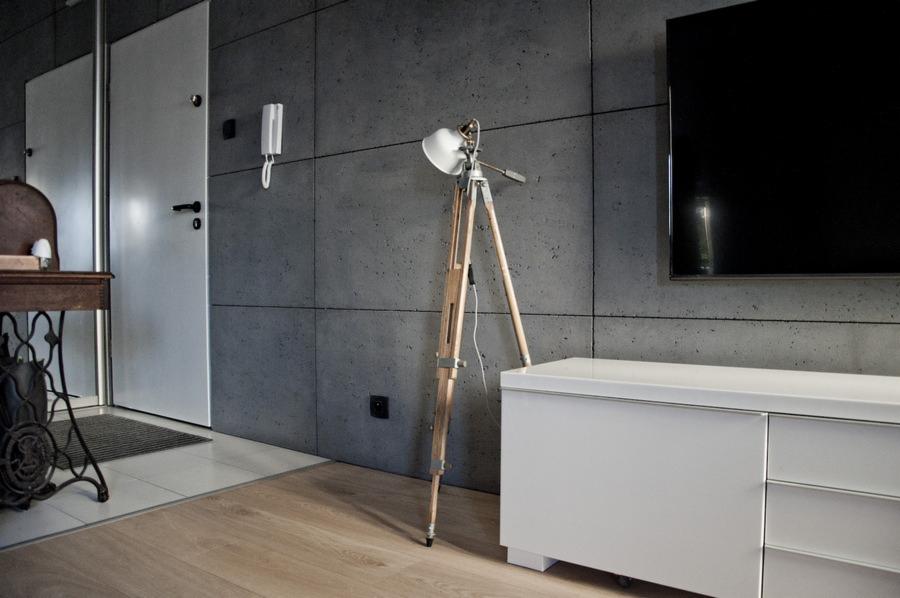 Wnętrze z betonem architektonicznym na ścianie, oświetlone lampą na trójnogu, z widoczną podłogą z jasnego drewna i elementami wystroju w stylu industrialnym.
