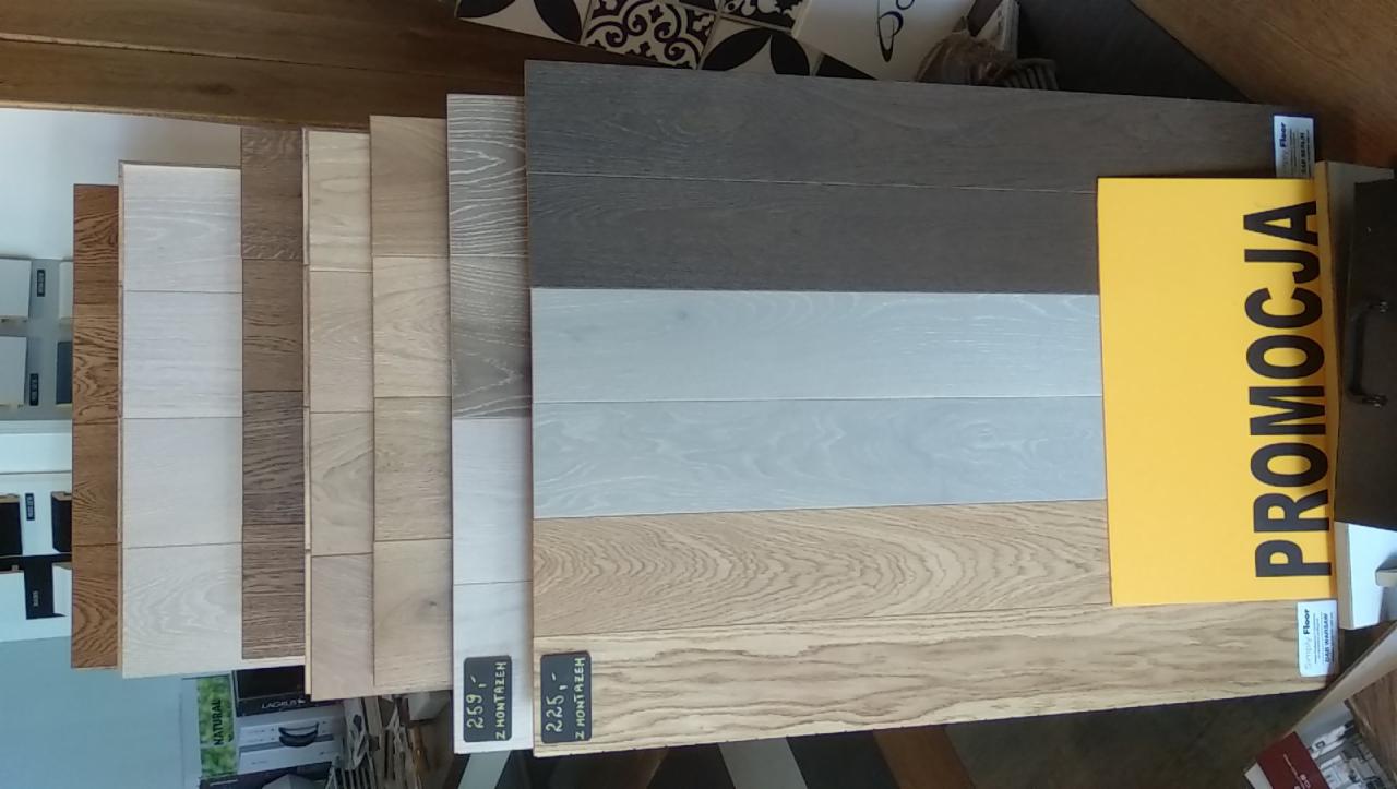 Ekspozycja próbek podłogowych w różnych odcieniach drewna, z żółtą kartką z napisem 'PROMOCJA'. Prezentacja materiałów wykończeniowych.