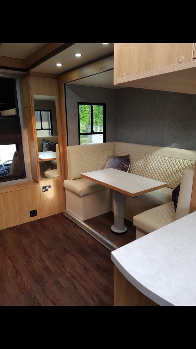 Jasne, pikowane obicie narożnej kanapy w kamperze, z drewnianymi elementami wykończenia i blatem stołu o jasnym odcieniu.