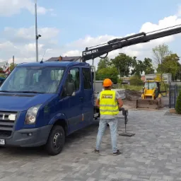 Niebieski samochód dostawczy z dźwigiem Hird 026 T, pracownik w kamizelce odblaskowej i kasku obsługuje urządzenie, w tle żółta koparka i kontener.