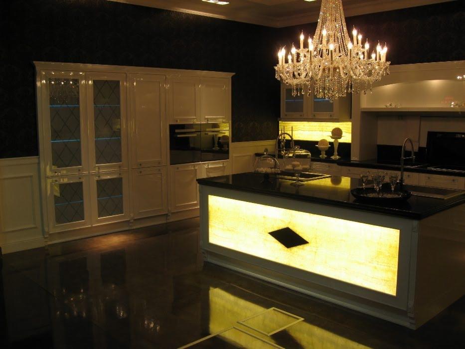 Elegancka kuchnia z białymi meblami w stylu klasycznym, oświetlona kryształowym żyrandolem, z wyspą z podświetlanym panelem i czarnym blatem, odbijająca się w polerowanej podłodze.