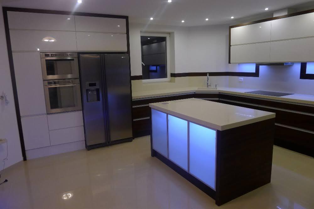 Nowoczesna kuchnia z wyspą oświetloną niebieskim światłem, białe szafki górne z ciemnobrązowymi obramowaniami, sprzęt AGD w zabudowie, jasna podłoga z połyskiem.