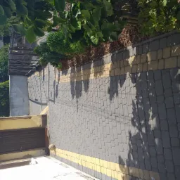 Ułożona szara kostka brukowa z żółtymi akcentami, obok zieleni i elewacji budynku z ciemnymi drzwiami.