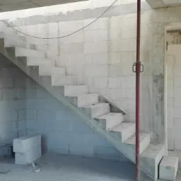 Surowe betonowe schody wewnętrzne w trakcie budowy, widoczne bloczki z betonu komórkowego, podpora stropowa z czerwonej stali i deska oparta o ścianę.