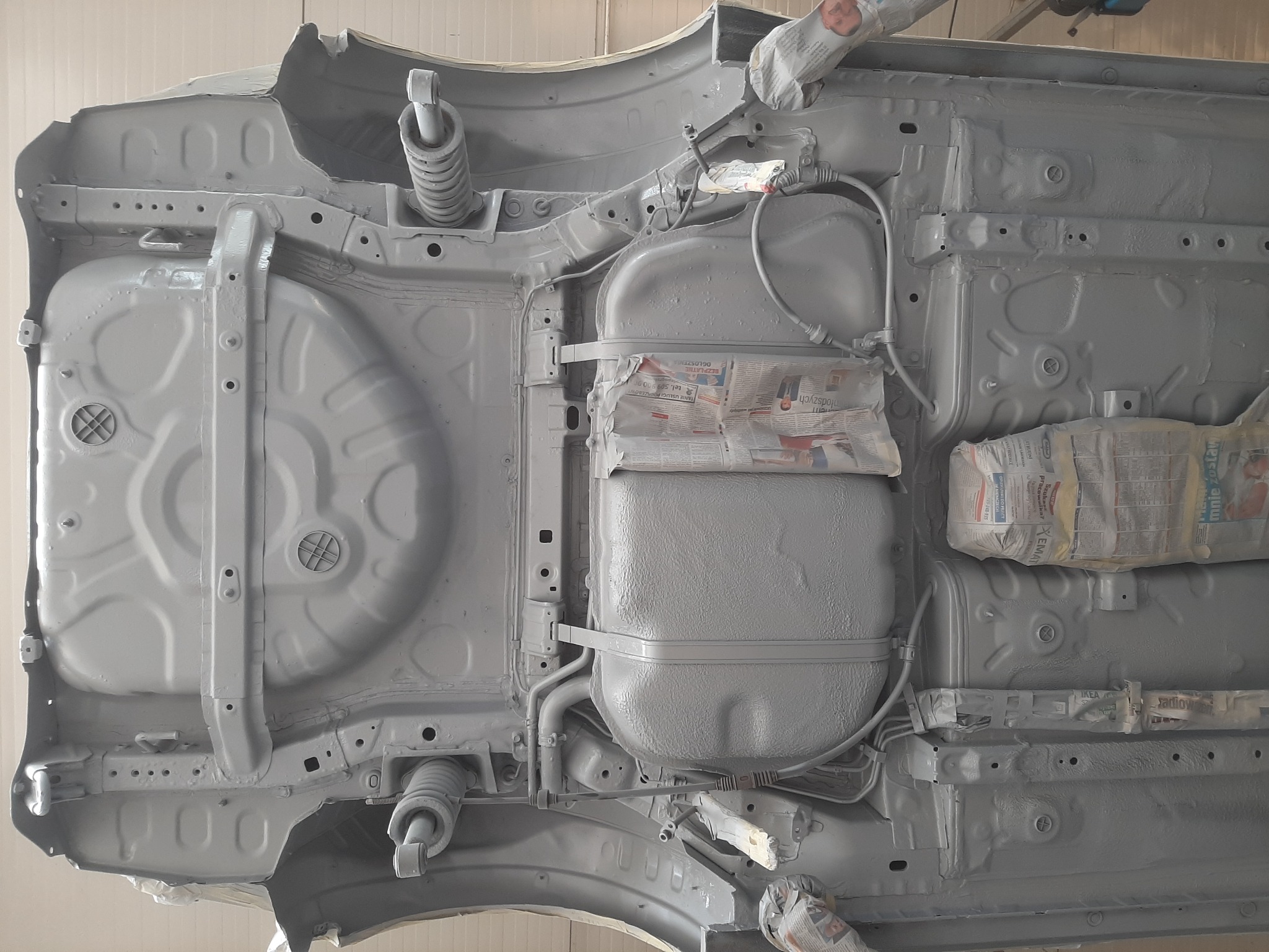 Spód samochodu po zabezpieczeniu antykorozyjnym, z widocznymi elementami zawieszenia i zbiornikiem paliwa osłoniętymi gazetami podczas malowania.