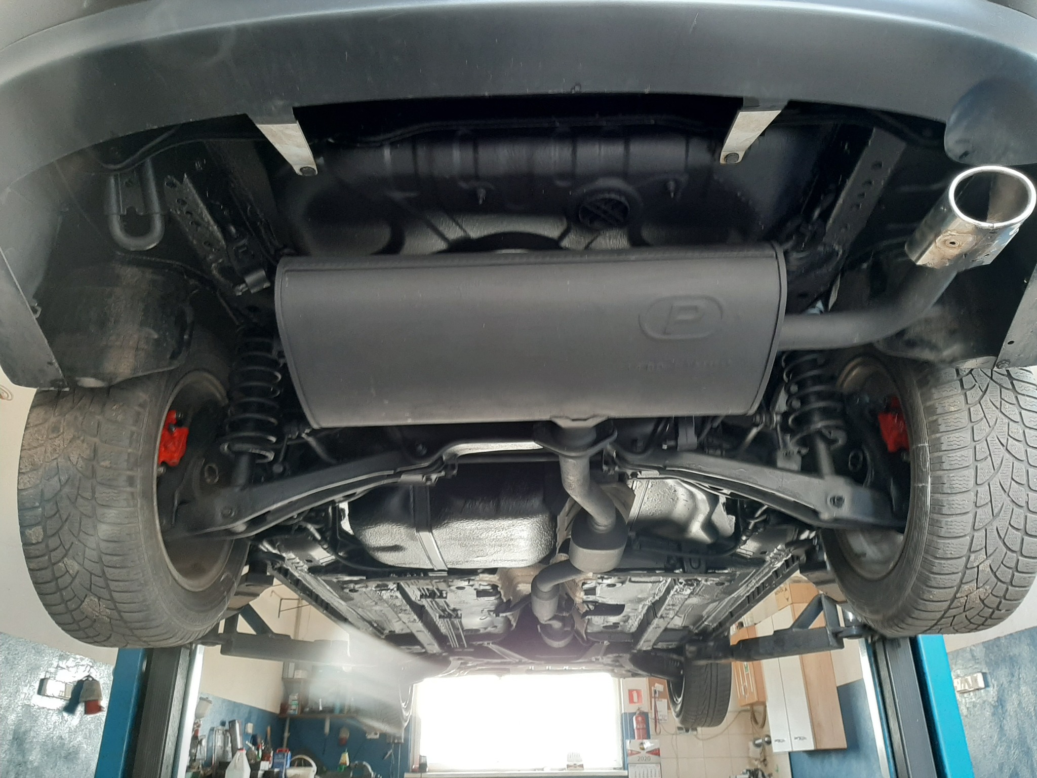 Podwozie samochodu osobowego po konserwacji antykorozyjnej, widoczne elementy zawieszenia, układ wydechowy z tłumikiem, felgi z oponami zimowymi, czerwone zaciski hamulcowe, całość podniesiona...