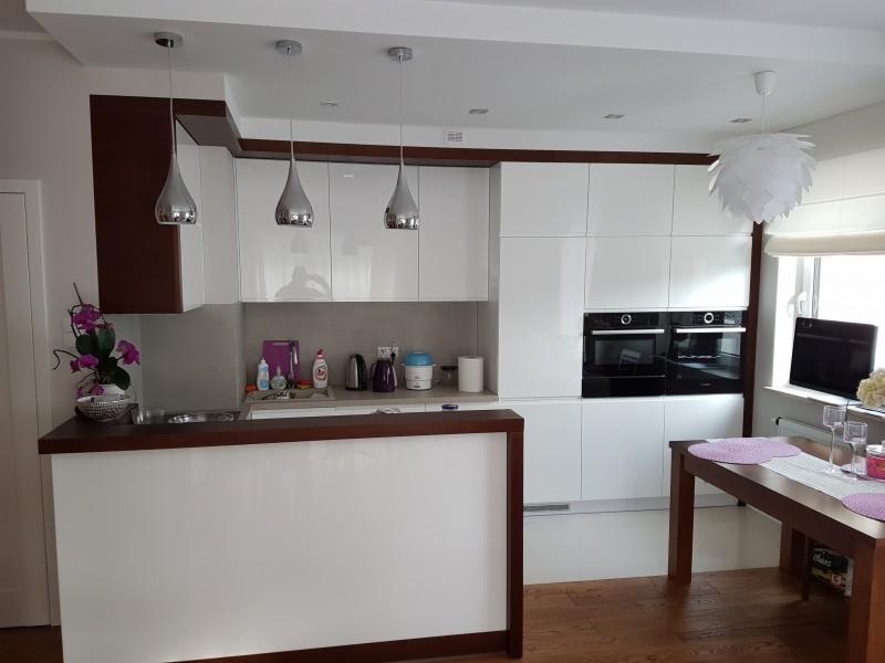 Nowoczesna kuchnia z białymi lakierowanymi frontami, ciemnobrązowymi akcentami, wyspą i trzema chromowanymi lampami wiszącymi nad blatem oraz dwoma piekarnikami w zabudowie.