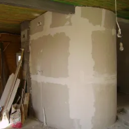 Niedokończony kominek o zaokrąglonym kształcie, częściowo pokryty gładzią, z widocznymi elementami konstrukcyjnymi i prowizorycznym oświetleniem żarówką na kablu.