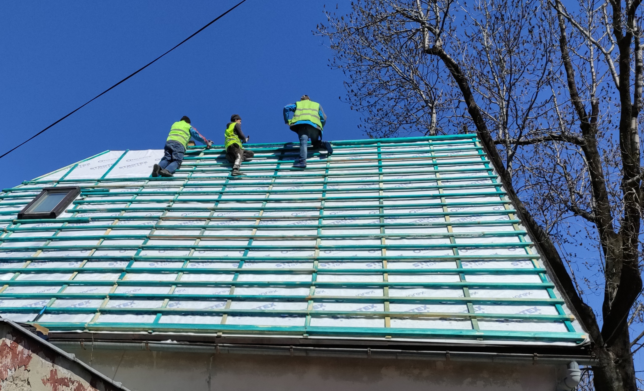 Trzech pracowników w kamizelkach odblaskowych montuje konstrukcję dachu z oknem dachowym, widok z dołu na tle błękitnego nieba i gałęzi drzewa.