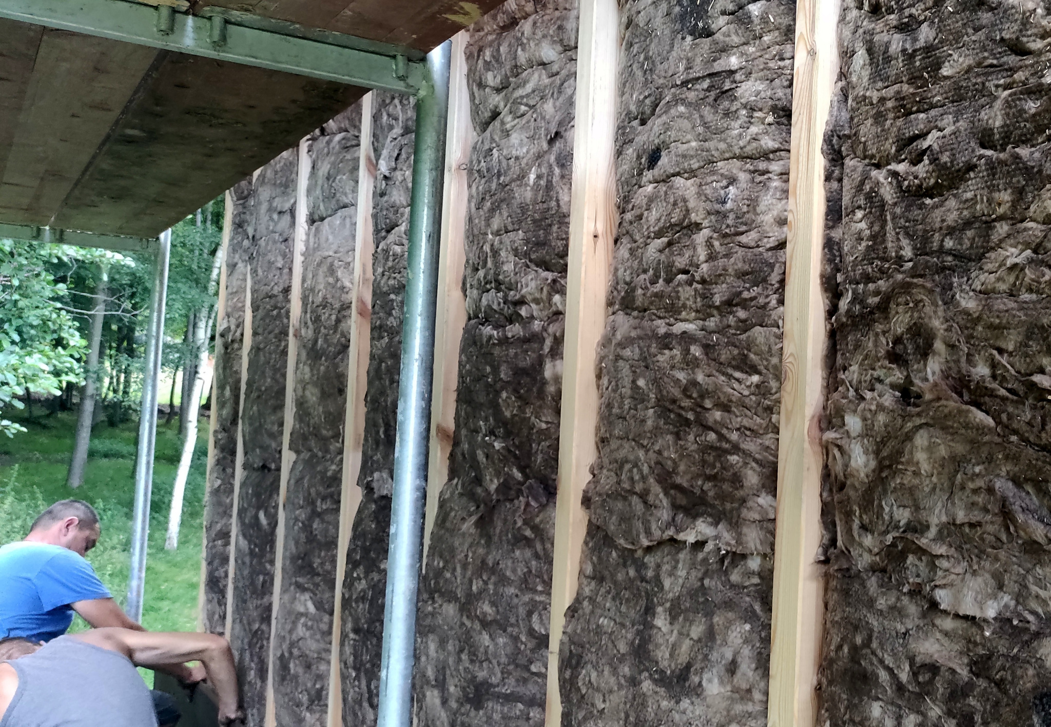 Montaż izolacji z wełny mineralnej w konstrukcji szkieletowej domu, widoczni pracownicy w trakcie pracy na zewnątrz budynku.