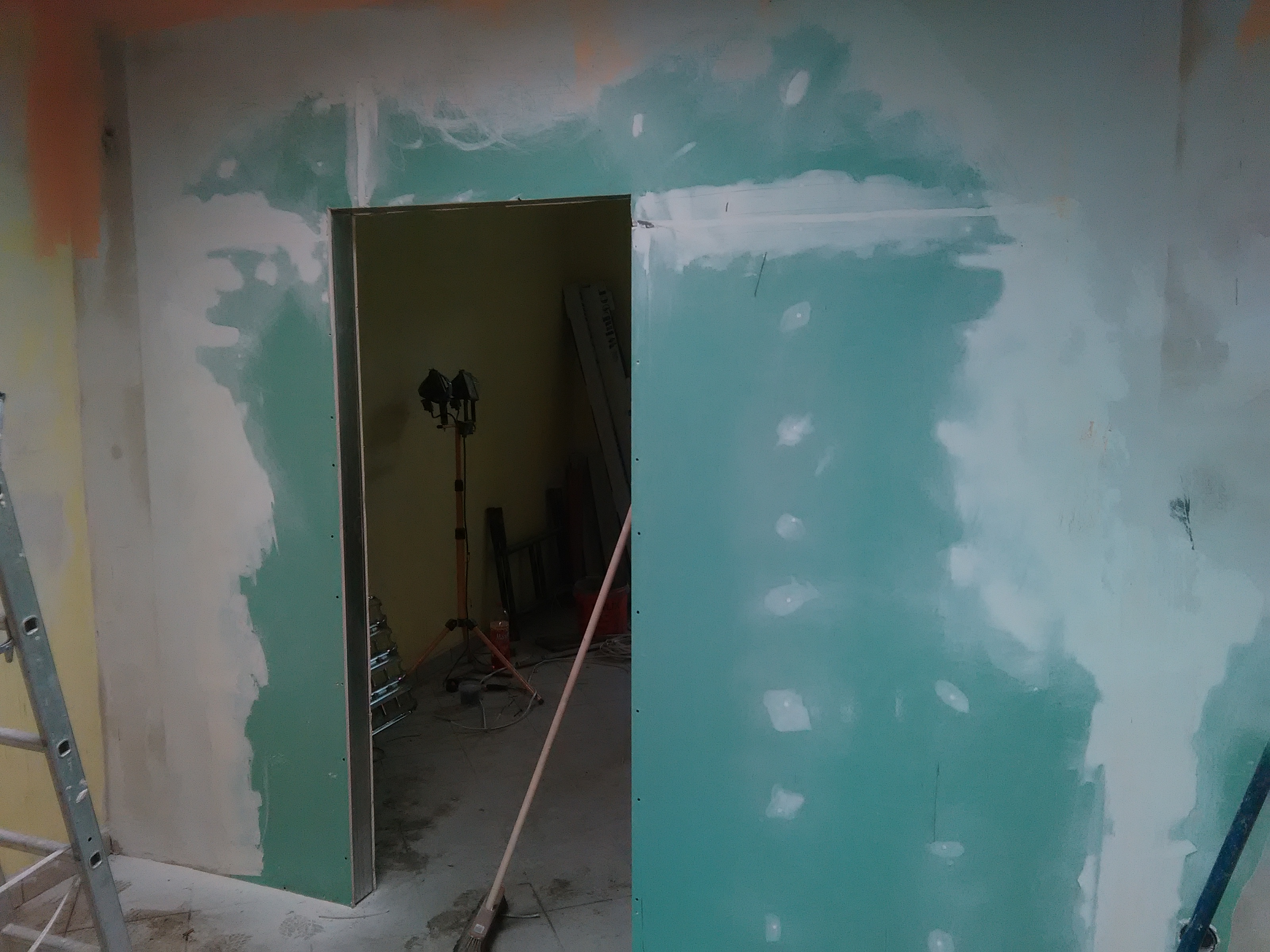 Wnętrze w trakcie wykańczania: ściany z płyt gipsowo-kartonowych, szpachlowane, widoczne ościeżnice drzwiowe, drabina i lampa budowlana.