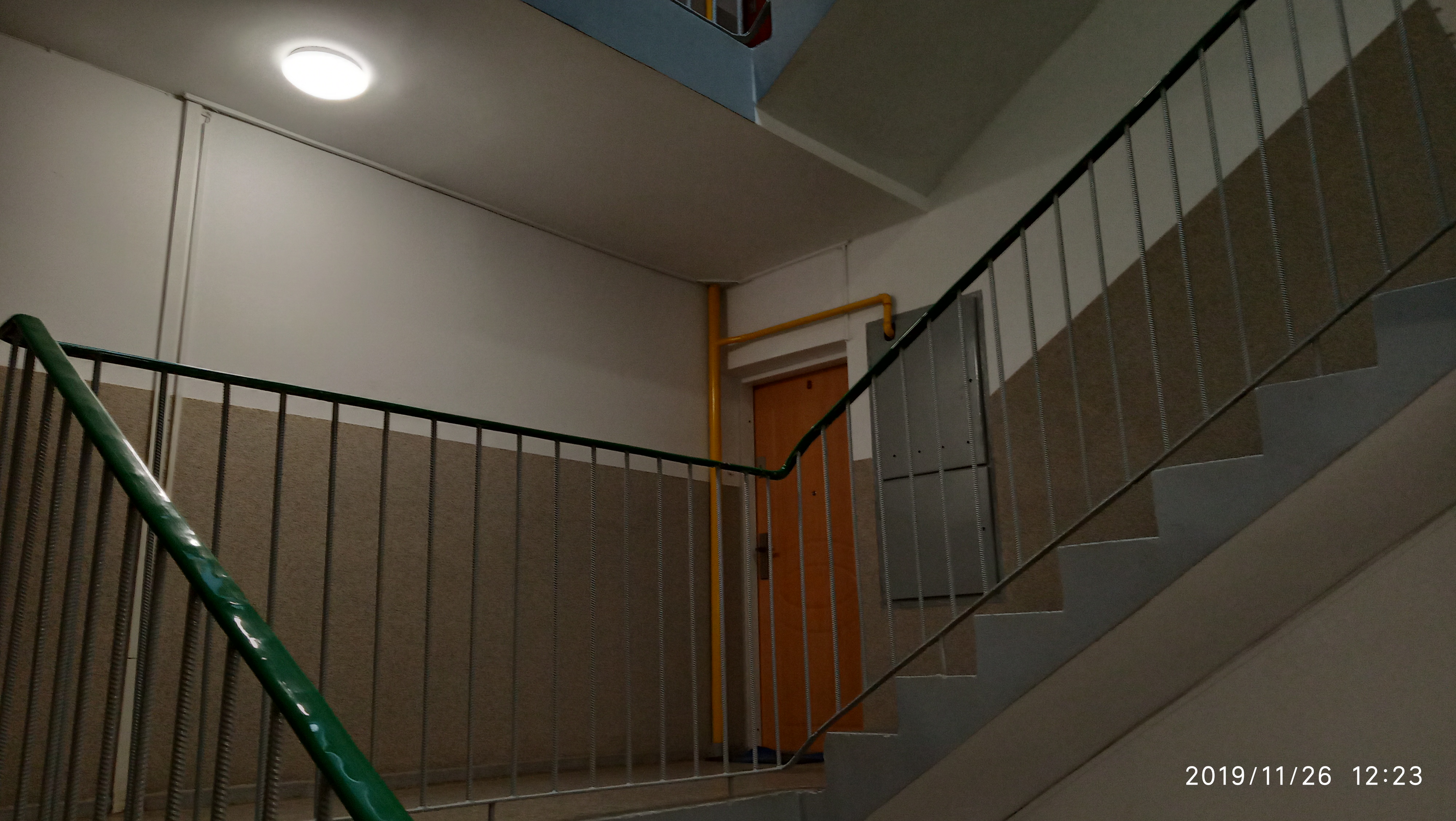 Wnętrze klatki schodowej z zieloną barierką, widoczne schody, drzwi i żółta rura instalacyjna na ścianie.