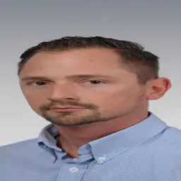 Portret mężczyzny w błękitnej koszuli z krótką brodą i ciemnymi włosami na jasnym tle.